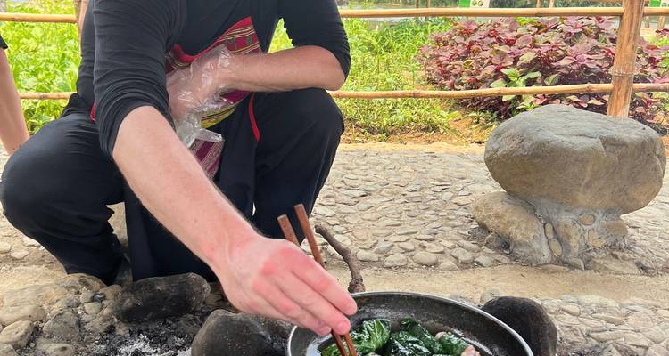 Close-up of a person stirring greens in a pot over an outdoor fire with chopsticks.