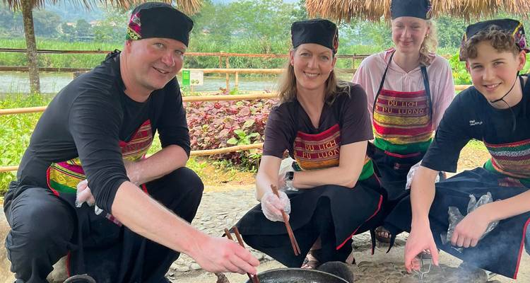 Four family members crouch around a cooking fire preparing food together outdoors.
