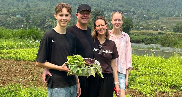 Family stands in a vegetable garden holding freshly picked greens with forested hills behind.
