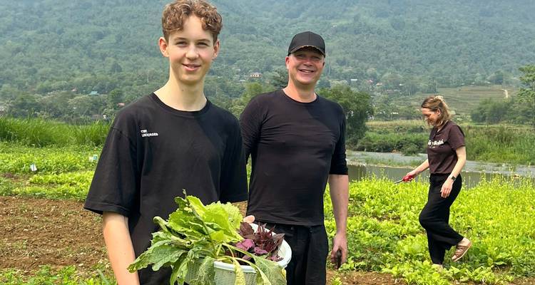 A teenage boy holds a basket of freshly picked greens beside two adults in a rural vegetable field with lush mountains in the background.