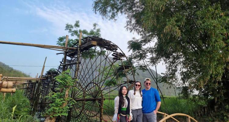 Three travelers pose beside a large bamboo waterwheel with green hills and bamboo groves behind them.