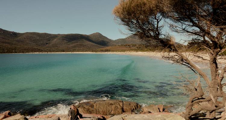Les eaux turquoise de Wineglass Bay rencontrant une plage de sable blanc encadrée par des collines escarpées et des arbustes.