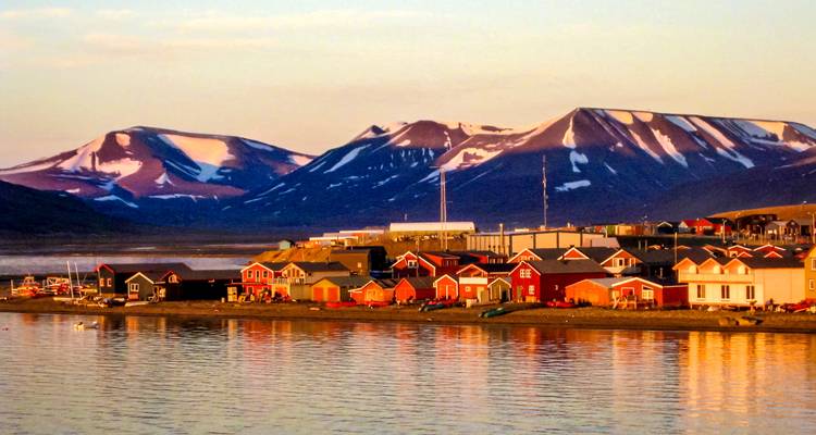 Warm evening light glows on the colorful coastal settlement of Longyearbyen with snow-tipped peaks behind a calm fjord.