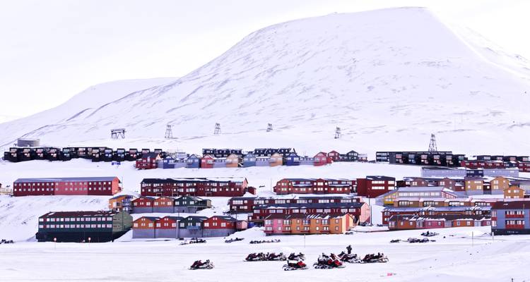 Winter panorama of Longyearbyen with red and brown buildings and dozens of snowmobiles parked on the frozen fjord beneath a towering white mountain.
