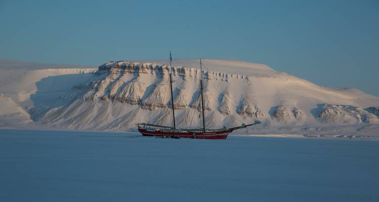 A red-hulled sailing ship frozen into an icy fjord below sheer snow-covered cliffs at dusk.