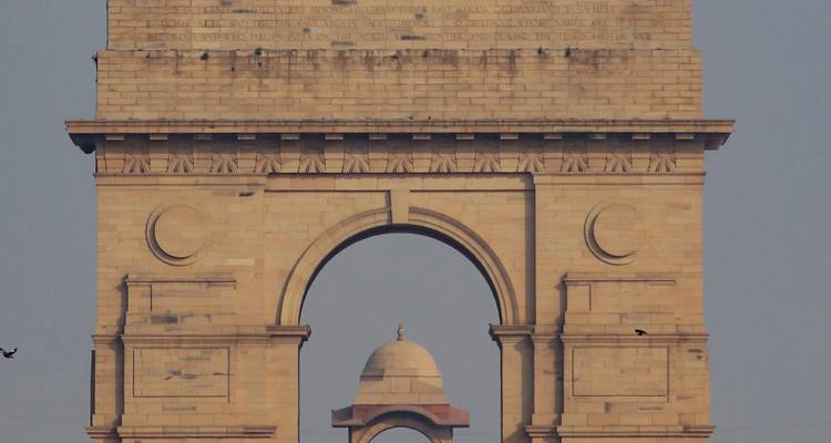 Overdag close-up van de zandstenen boog en koepel van India Gate tegen een lichtblauwe hemel.