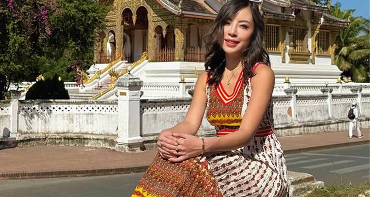 Vrouw in een gedessineerde jurk zit glimlachend voor een prachtige goud en witte tempel in Luang Prabang.