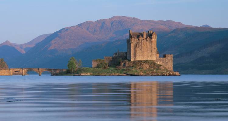 Eilean Donan Castle op een klein eilandje weerspiegeld in stille loch tegen ruige bergachtergrond bij schemering.