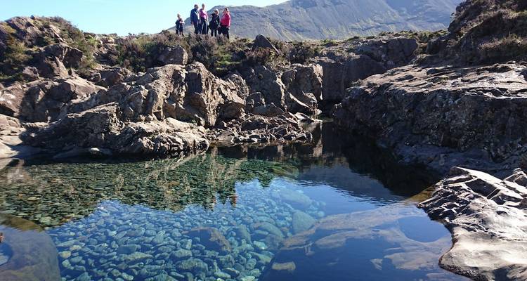 Kristalhelder rotsbadje van de Fairy Pools met wandelaars die op de rotsige rand staan en het Cuillin gebergte daarachter.