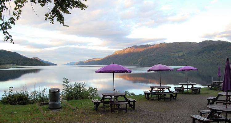 Picknicktafels met paarse parasols naast een rustig Hooglanden meer in het gouden zonlicht.