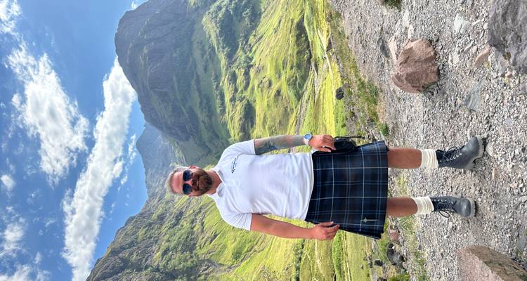 Rondleidingsgids in kilt poserende in dramatische bergvallei van Glencoe onder blauwe lucht.