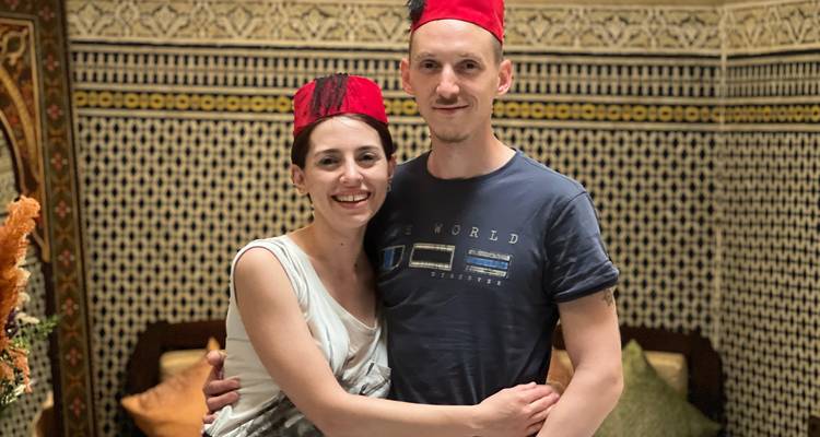 Couple souriant portant des chapeaux de fez rouges à l'intérieur d'un intérieur marocain richement carrelé.