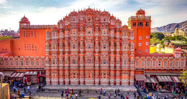Lebendige Fassade des Hawa Mahal Palastes in Jaipur mit Menschenmengen und Marktständen, die sich entlang der Straße unten aufgereiht haben.