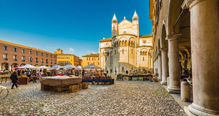 Openluchtantieksmarkt op Modena's kasseienplein omgeven door galerijen met de kathedraal die gloeït in het late middaglicht.