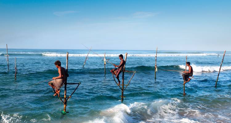 Trois pêcheurs sri-lankais en équilibre sur des échasses en bois au-dessus de vagues bleu clair avec de l'écume blanche en arrière-plan.