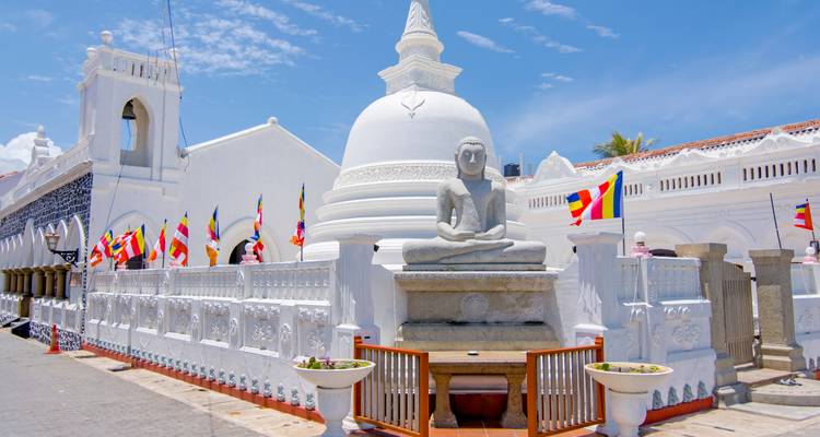 Un dagoba blanc serein avec une statue de Bouddha assis est entouré de drapeaux de prière colorés et de balustrades ornées sous un ciel bleu éclatant.