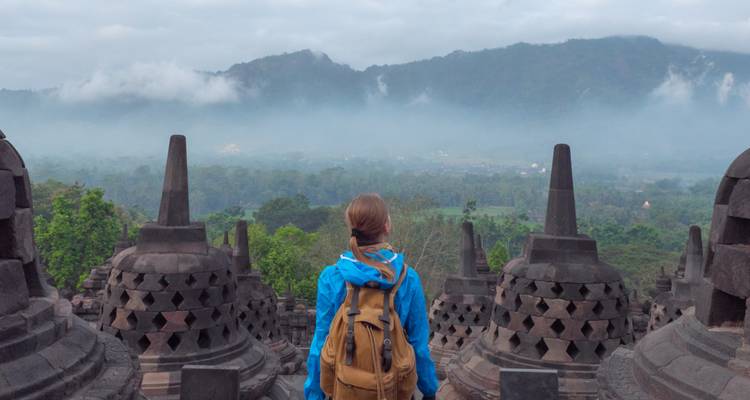 A traveler in a blue jacket looks over misty jungles from the stone stupas of Borobudur.