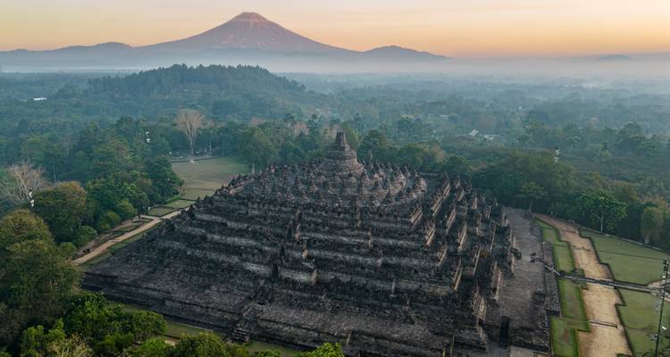 Aerial sunrise over Borobudur temple complex with Mount Merapi silhouetted in the distance.