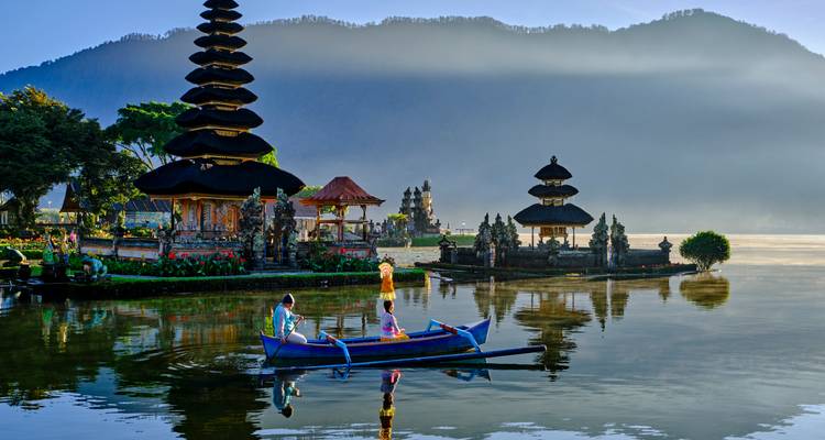 Pequeña canoa azul con turistas remando en un lago tranquilo junto al templo Pura Ulun Danu Bratan envuelto en niebla al amanecer.
