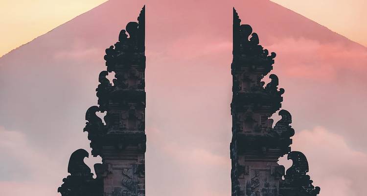Nahaufnahme einer Silhouette eines balinesischen Tempeltors, das sich vor einem nebligen Vulkangipfel gegen einen pastellfarbenen Sonnenuntergangshimmel abzeichnet.