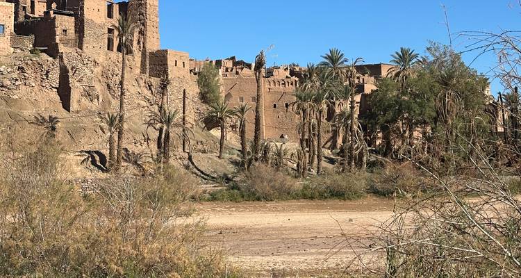 Earthen kasbah ruins and palm trees lining dry riverbed under deep blue sky.