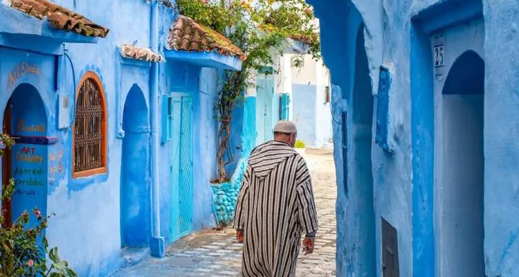 Ein traditionell gekleideter Mann geht eine enge blaue Gasse in Chefchaouen hinunter.