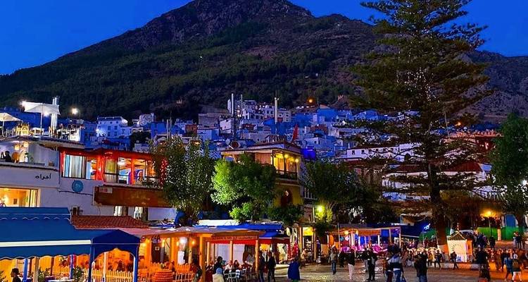 La place principale de Chefchaouen illuminée la nuit avec des cafés animés et des habitants sous un décor montagneux.