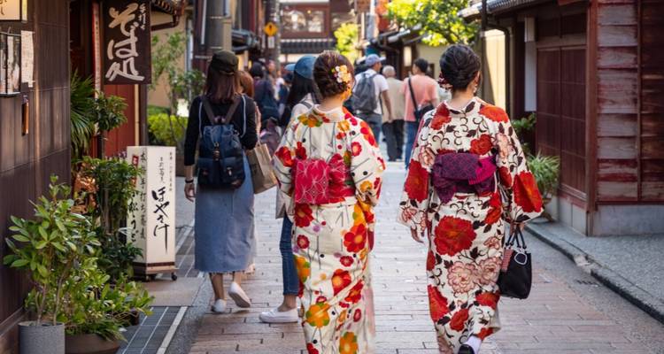 Zwei Frauen in blumigen Kimonos, die eine traditionelle Gasse mit Holzgeschaeften entlanggehen