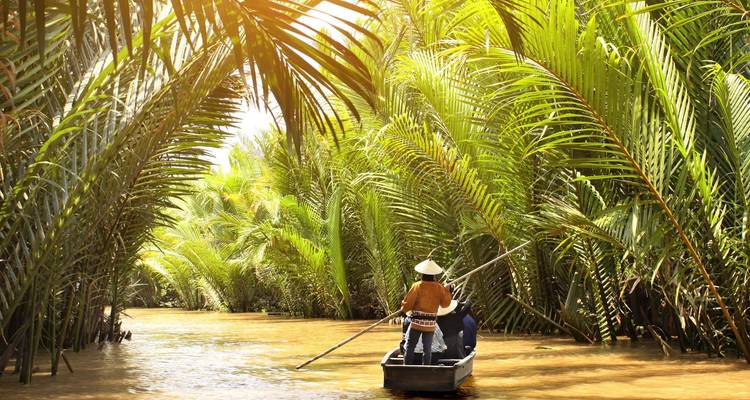 Ein kleines Sampan navigiert einen schmalen, mit Palmen bedeckten Wasserweg, der von warmer Sonneneinstrahlung durchflutet wird, im Mekong-Delta.