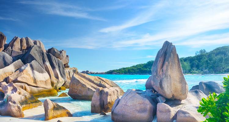 Kristallklares blaues Meer, das gegen glatte Granitfelsformationen unter einem strahlend blauen Himmel an einem tropischen Strand brandet.