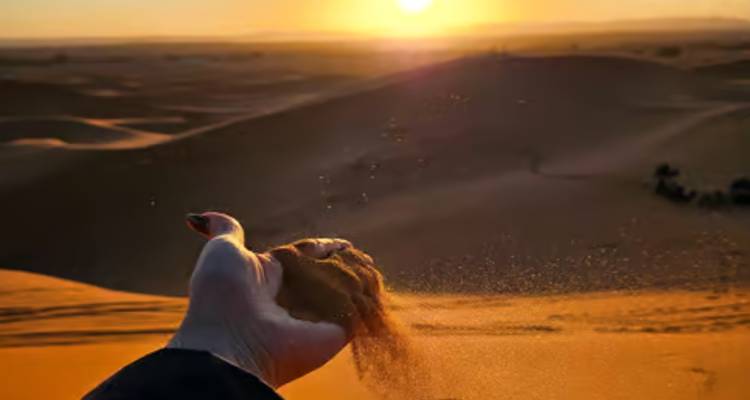 Une main laissant tomber le sable à travers les doigts sur une dune tandis que le soleil se couche sur le désert vallonné