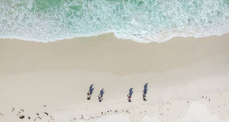 Vue aérienne d'une plage de sable blanc immaculé avec des vagues turquoise et cinq randonneurs projetant de longues ombres