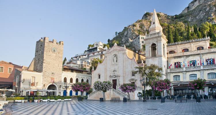 Charmante hoofdplein van Taormina met historische kerk, klokkentoren, bloeiende bomen en omringende heuvelhuis huizen onder een heldere hemel.