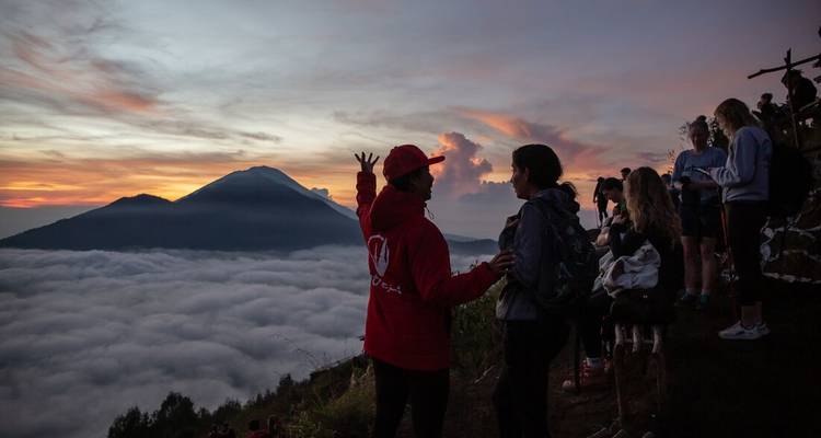 Group of hikers on a mountain ridge at sunrise overlooking cloud-filled valley and volcanic peak.