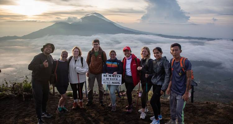 Happy trekking group poses on volcanic summit above a sea of clouds with a mountain backdrop.