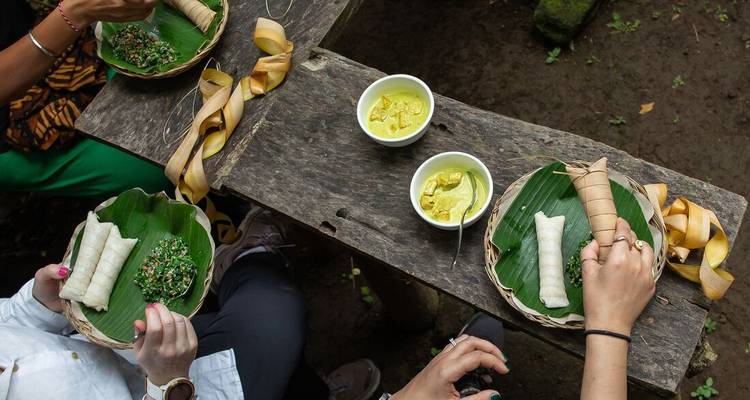 Top-down view of diners sampling Balinese dishes wrapped in banana leaves at a rustic wooden table.