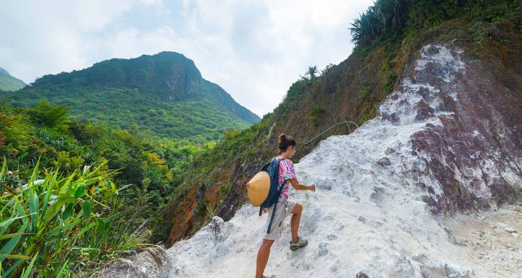 Backpacker climbs a rocky sulphur trail on a lush volcanic mountainside carrying a large basket.