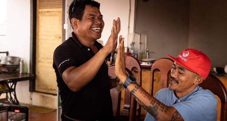 Two Cambodian men share a joyful high-five inside a modest home setting.