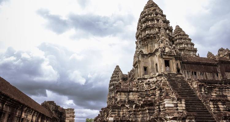 Angkor Wat stone towers rise against a dramatic cloudy sky, captured from a side angle.