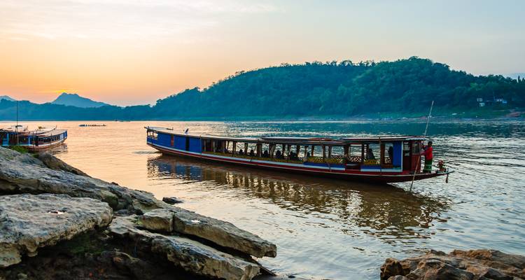 A long wooden riverboat floats on calm golden water at sunset with forested hills silhouetted behind.
