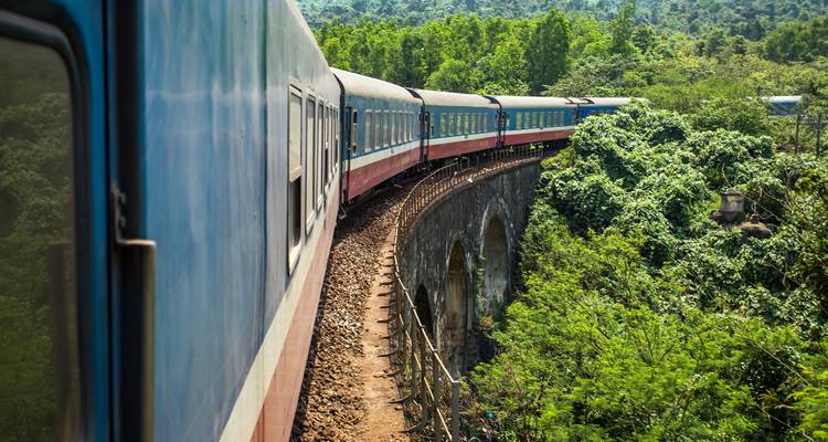A long blue passenger train curves across a high stone bridge through dense green hills.