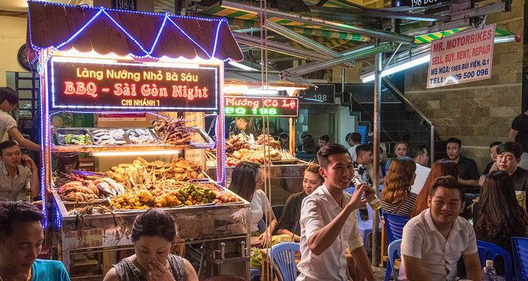 A vibrant night food market stall in Saigon advertises BBQ as diners eat and chat nearby.