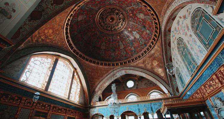 Interior de una sala del palacio otomano con una cúpula y arcos ricamente decorados en rojos y oros intensos.