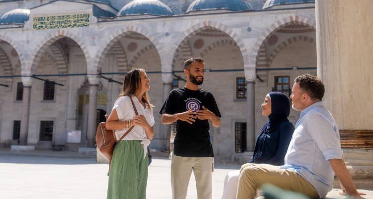 Guide explains history to travelers inside an ornate mosque courtyard with arched colonnades.