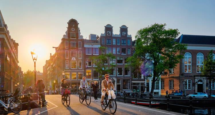 Cyclists ride across an Amsterdam canal bridge at sunset with lens flare lighting historic brick houses.