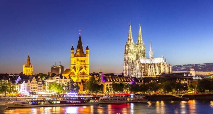 Twilight skyline of Cologne featuring the illuminated cathedral and historic churches reflected on the Rhine.