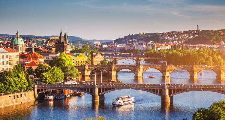 Plusieurs ponts historiques en pierre enjambent la rivière Vltava à Prague, avec un bateau de tourisme et un paysage urbain illuminé par le soleil dans la lueur de fin d'après-midi.