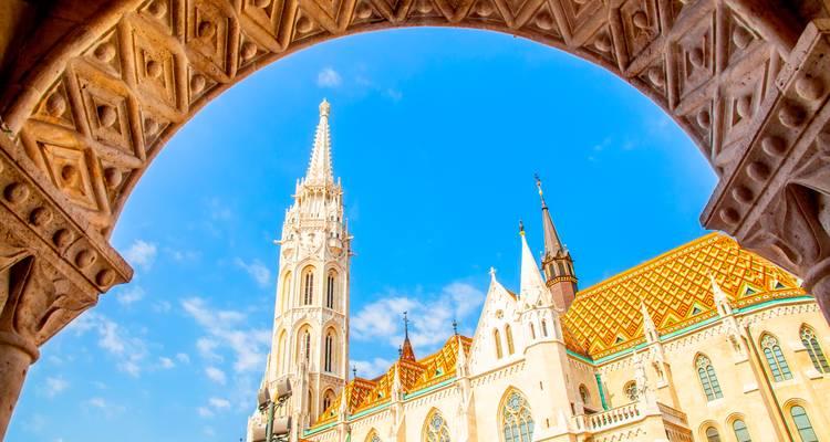 Bright view of Matthias Church framed by an ornate stone arch against a vivid blue sky in Budapest.