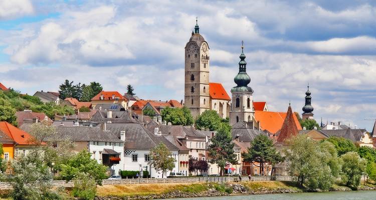 Riverside Austrian town with multiple church spires and colorful houses beneath a partly cloudy sky.