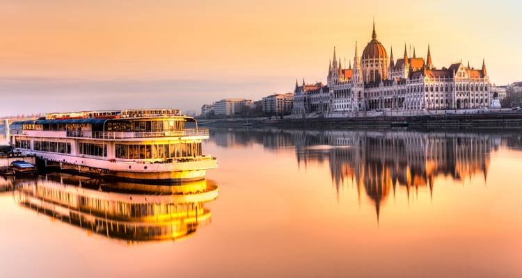 Reflet du lever de soleil doré d'un bateau de croisière fluviale et du Parlement hongrois sur les eaux calmes du Danube.